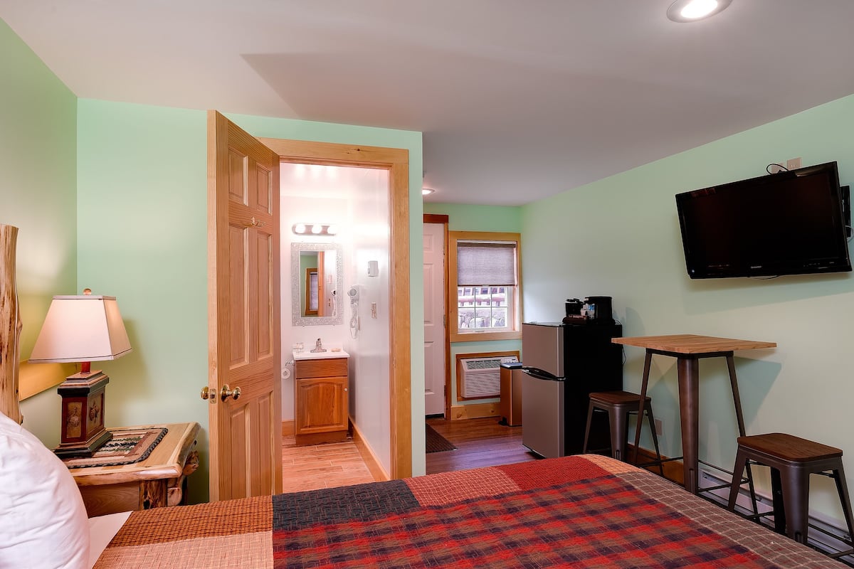 A bedroom with a bed, a TV mounted on the wall, a small table, and a refrigerator.