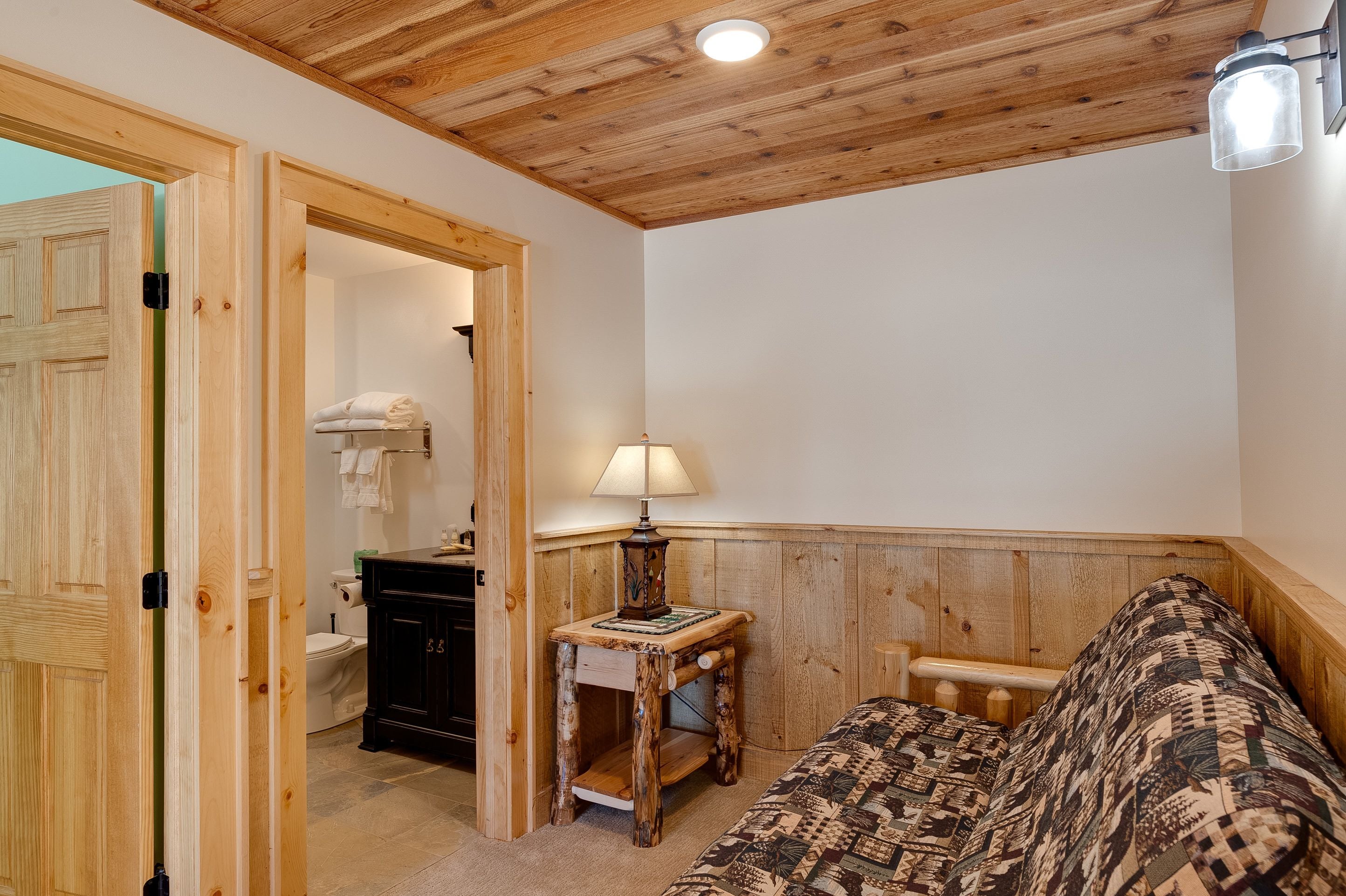 A room with a wooden ceiling, a bed with patterned bedding, a bedside table with a lamp, and a door leading to a bathroom.