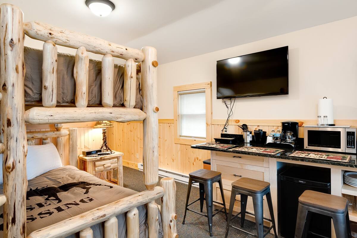 A rustic cabin-style room with a wooden bed, a television mounted on the wall, a kitchenette with a microwave, and a small bar area with stools.
