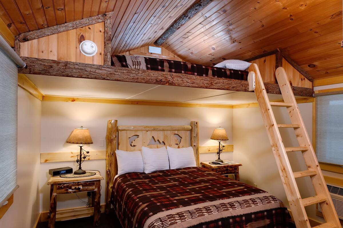 A cozy wooden cabin bedroom with a loft bed, two bedside tables with lamps, a ladder, and a large window.