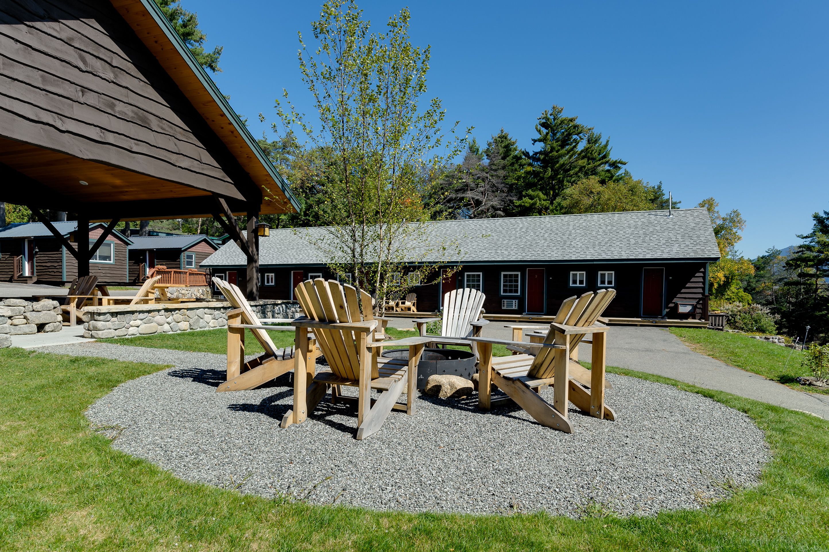 A wooden deck with Adirondack chairs and a table, surrounded by a gravel area with a building in the background.