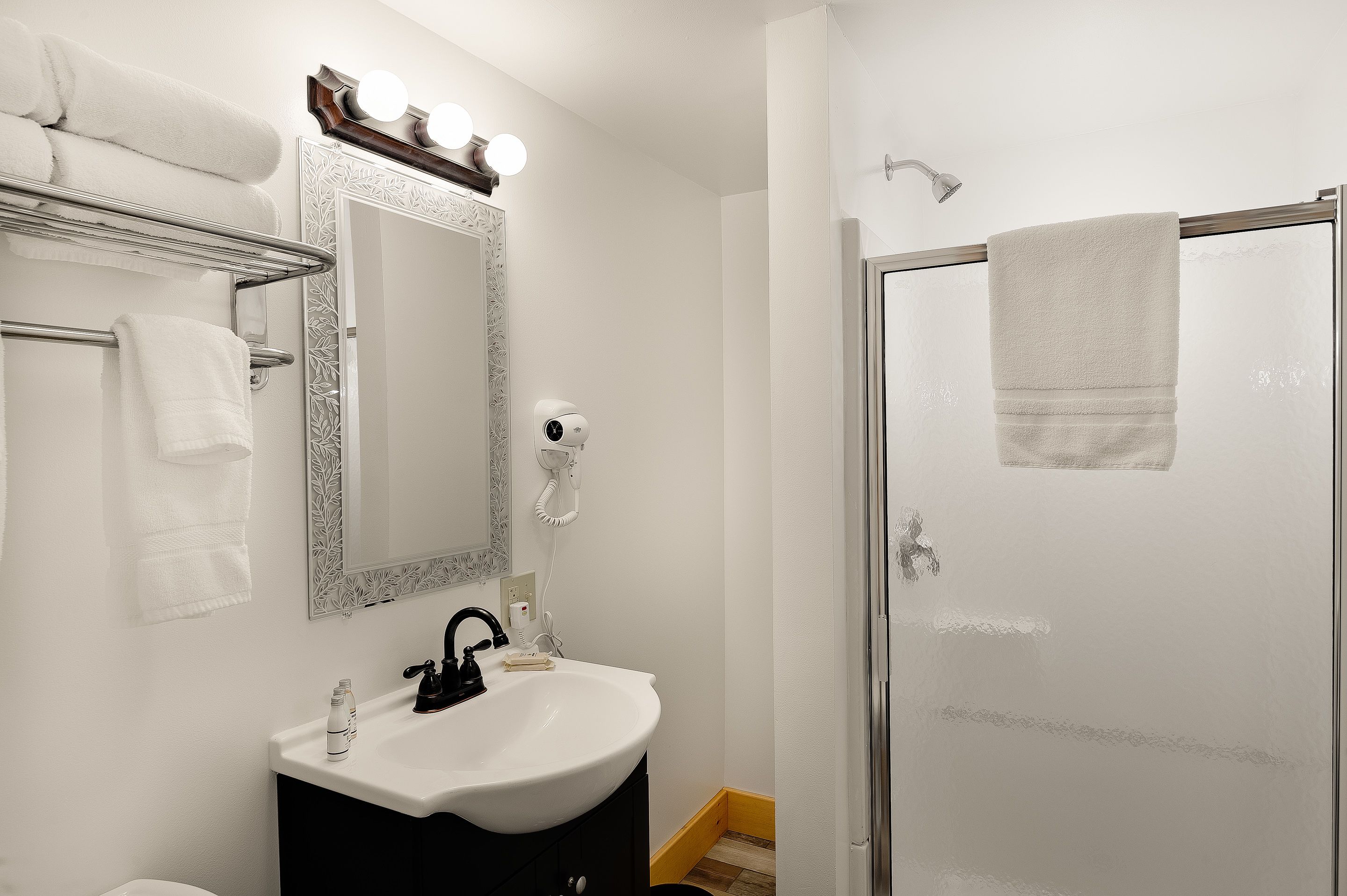 Casa de banho com lavatório branco, espelho com iluminação, secador de cabelo e cabine de duche em vidro.