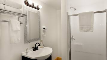 Casa de banho com lavatório branco, espelho com iluminação, secador de cabelo e cabine de duche em vidro.