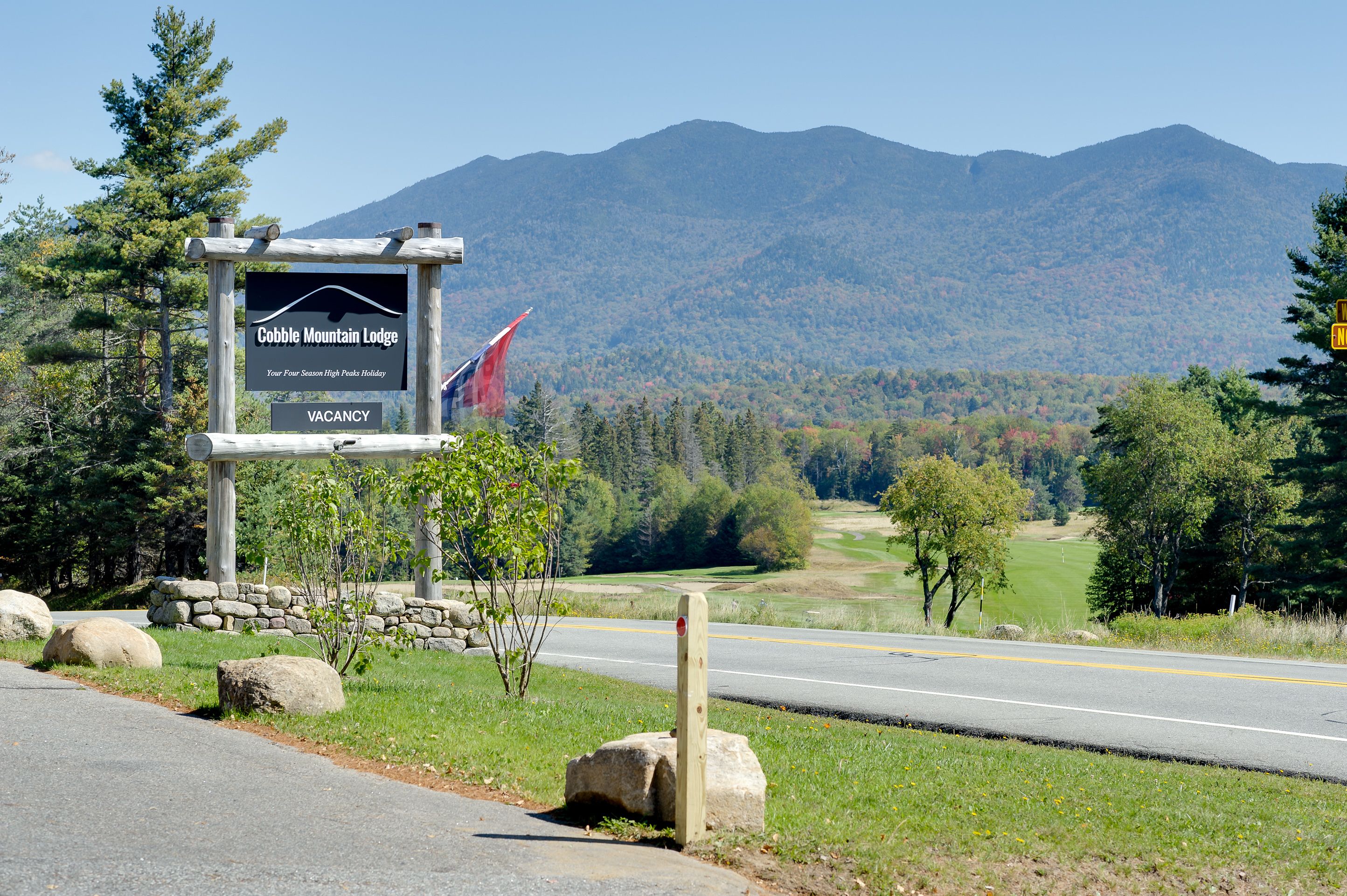 A sign for Cobble Mountain Lodge with a vacancy notice, set against a backdrop of mountains and a clear road.