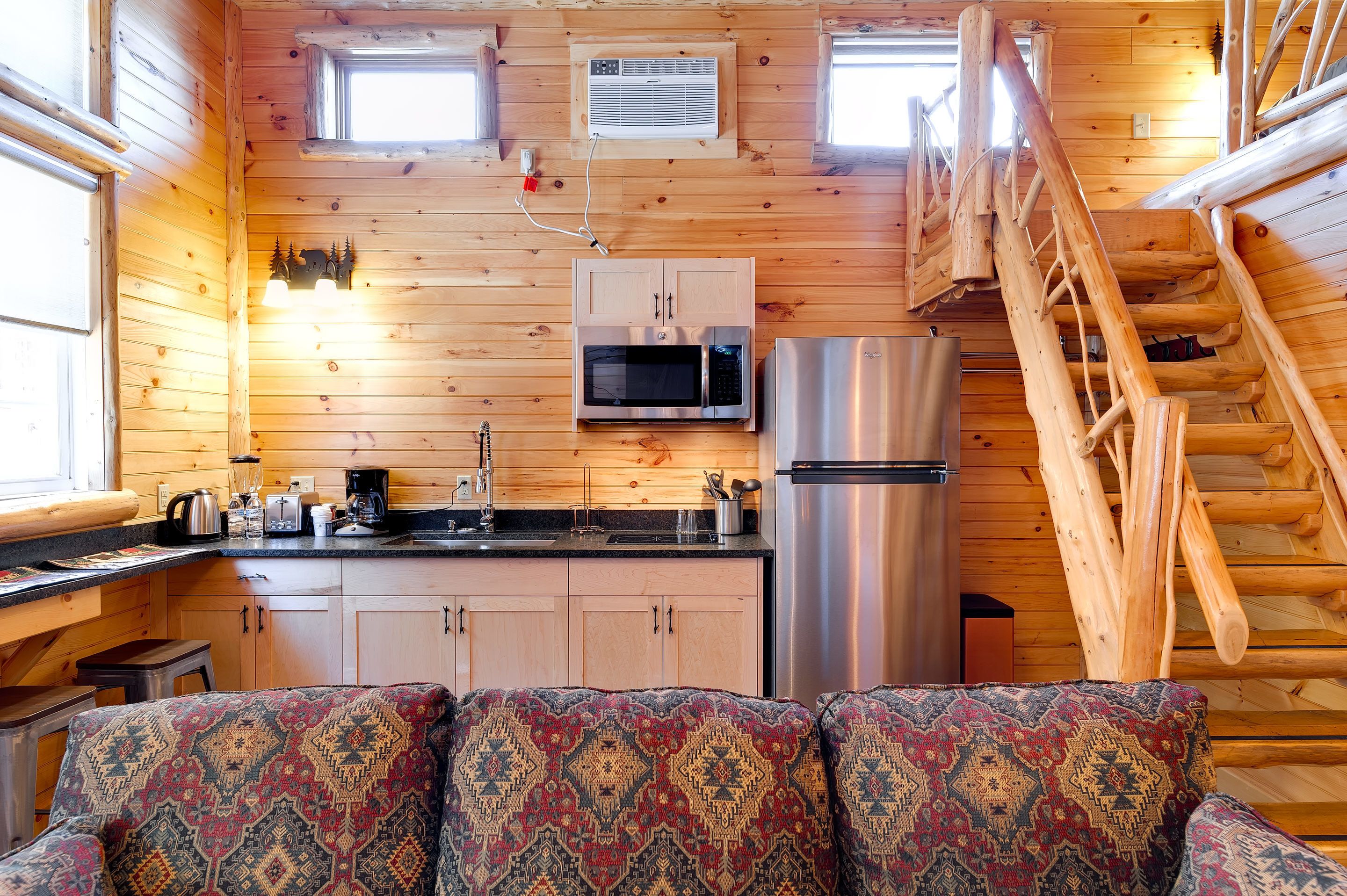 Un acogedor interior de cabaña con detalles de madera, una kitchenette con microondas y refrigerador, un sofá con cojines estampados y una escalera de madera.