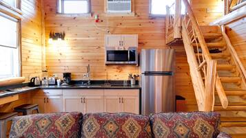 Un acogedor interior de cabaña con detalles de madera, una kitchenette con microondas y refrigerador, un sofá con cojines estampados y una escalera de madera.