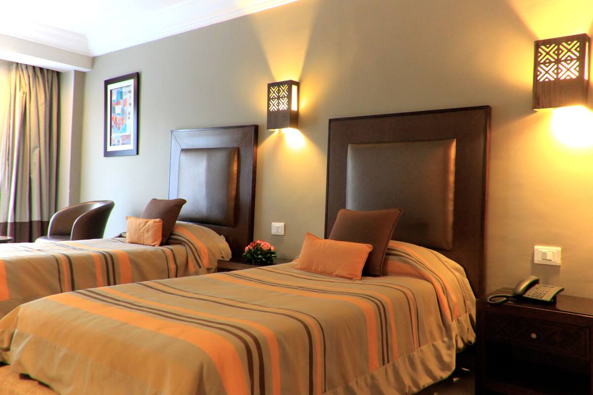 Habitación de hotel con dos camas, cada una con ropa de cama a rayas y cabeceros a juego. Se observan mesitas de noche con lámparas y un teléfono.