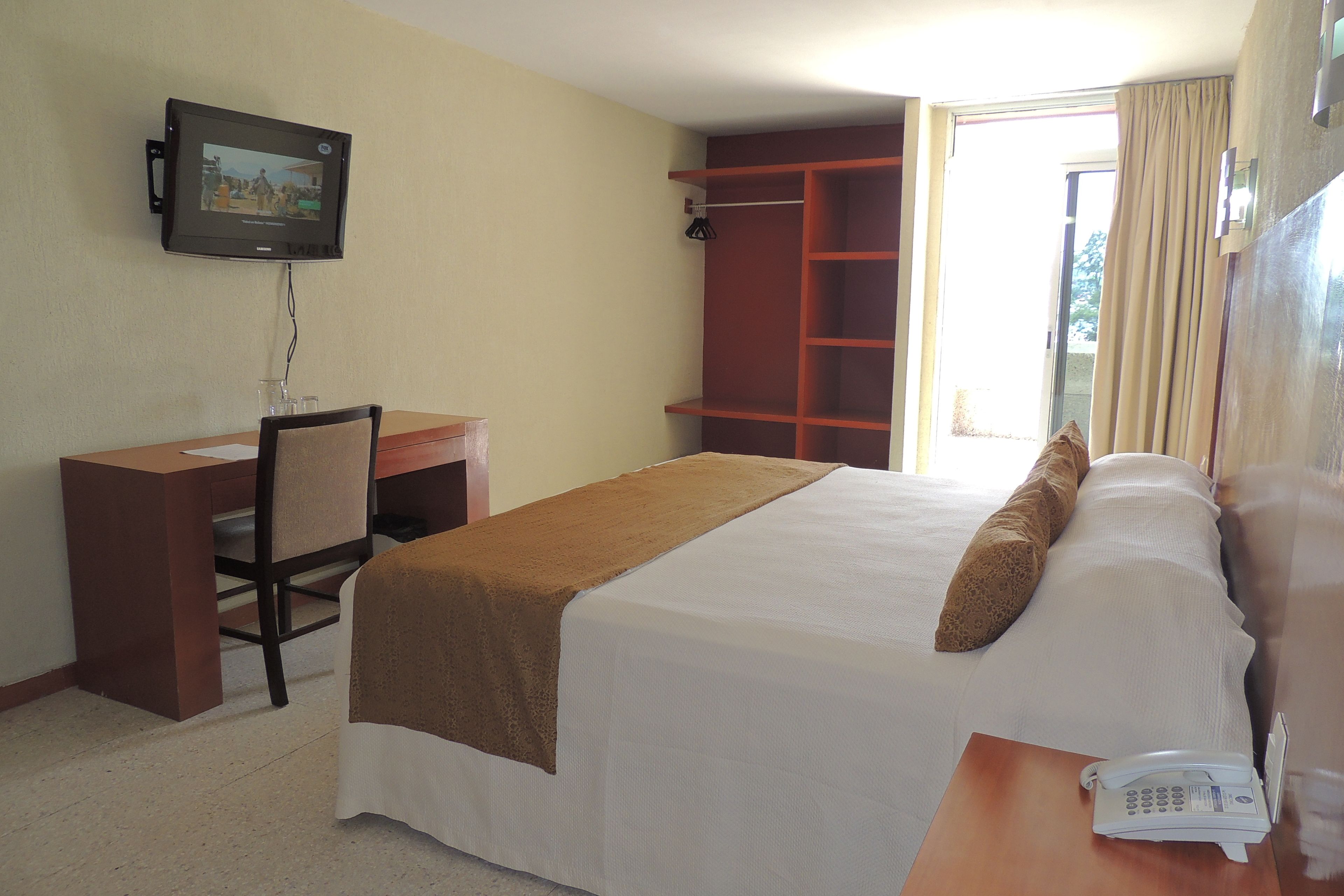 A hotel room with a bed, a desk, a chair, a TV mounted on the wall, and a telephone on the bedside table.