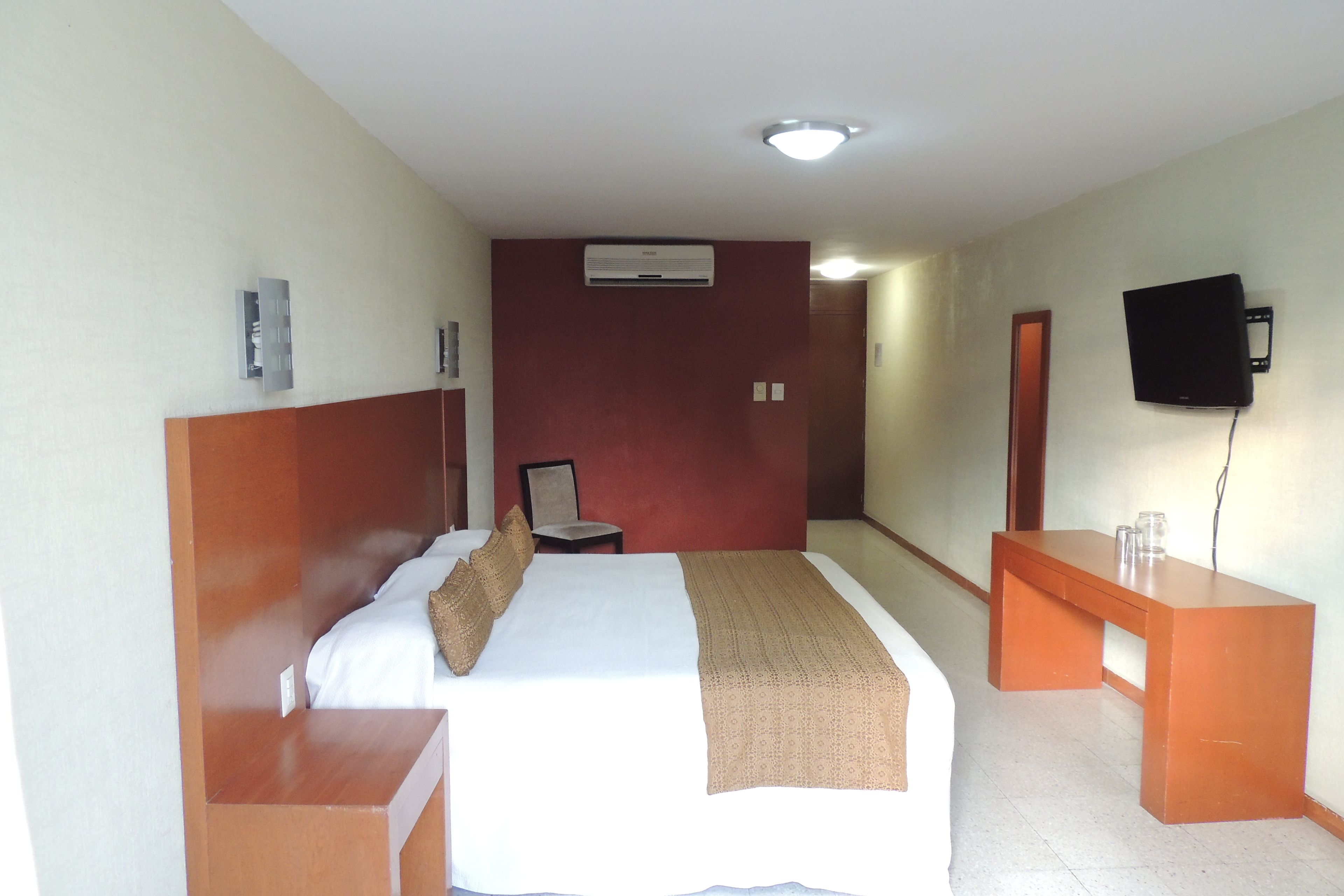 Una habitación de hotel con una cama, mesita de noche, escritorio, silla, televisor montado en la pared y equipo de aire acondicionado.