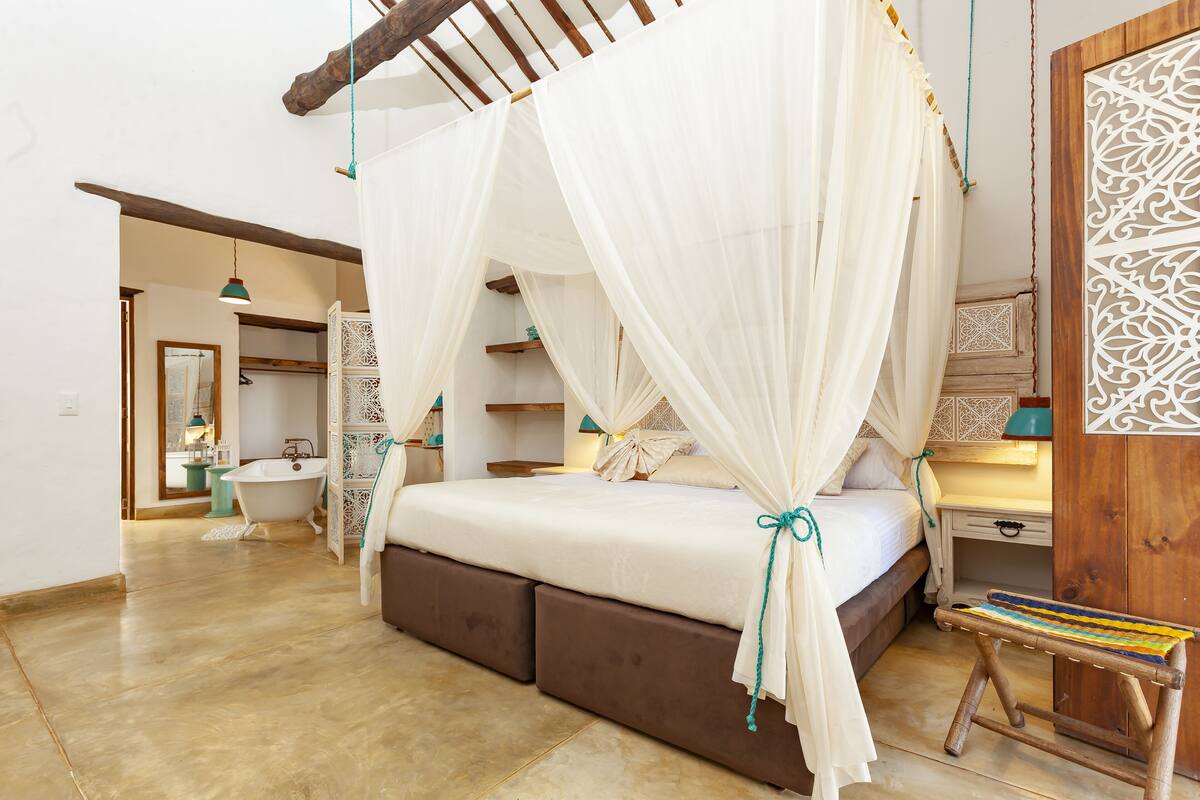A bedroom with a bed under a canopy, a wooden bench, a nightstand, and a bathroom with a bathtub and sink.