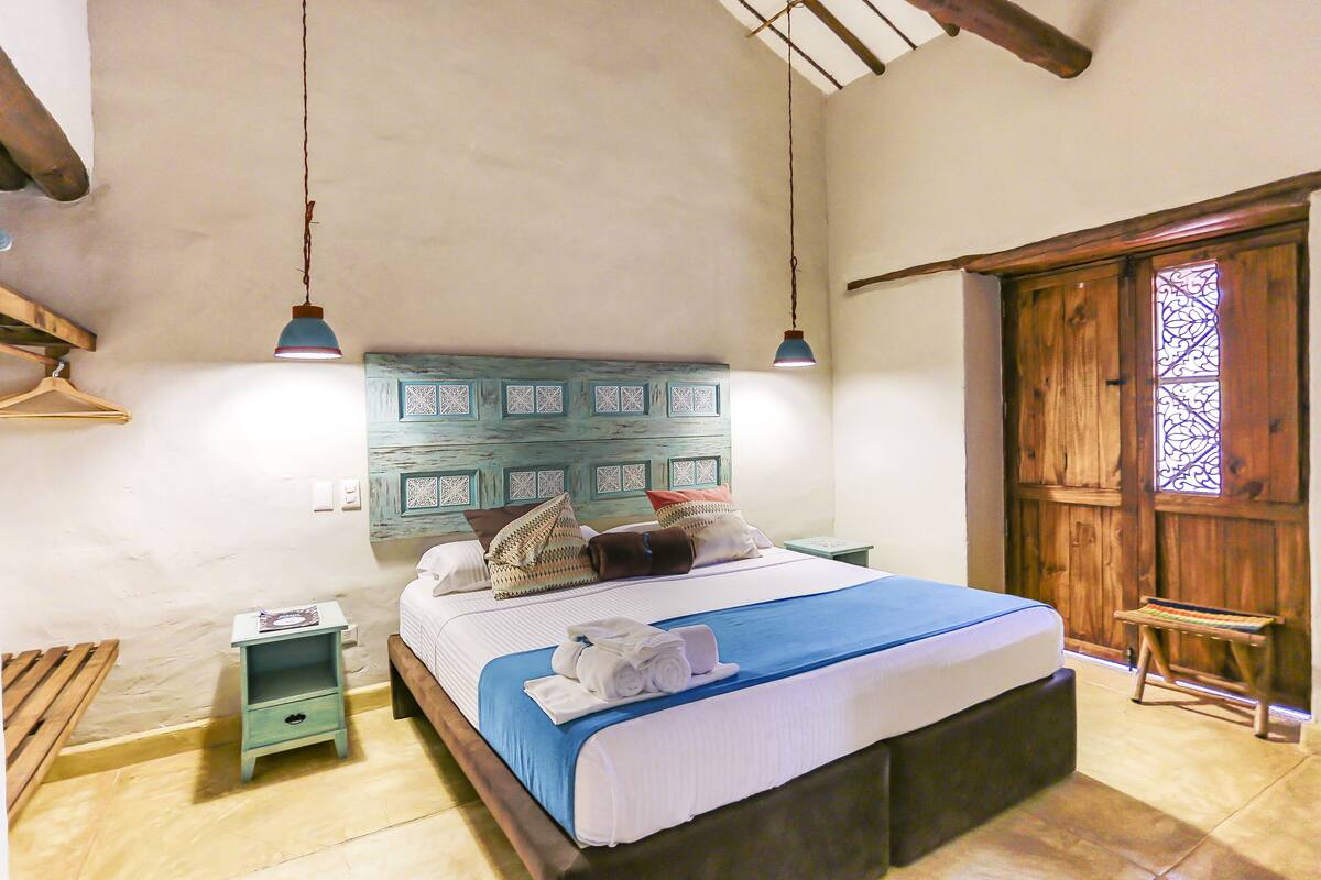 A bedroom with a wooden headboard, a bed with a blue bedspread, a wooden bench, and a small table with a lamp.