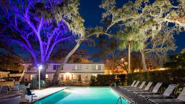 Una piscina con sillas de descanso, un edificio con un porche y árboles con iluminación morada.
