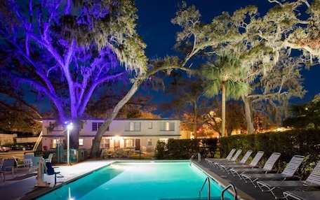 A swimming pool with lounge chairs, a building with a porch, and trees with purple lighting.