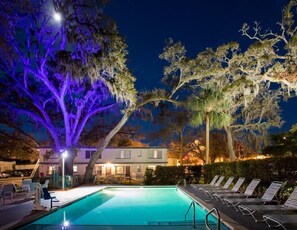 Una piscina con sillas de descanso, un edificio con un porche y árboles con iluminación morada.