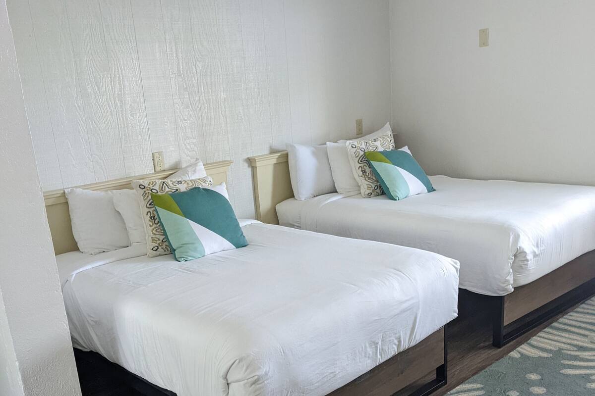 Two single beds with white linens and decorative pillows, a wooden floor, and a patterned rug.
