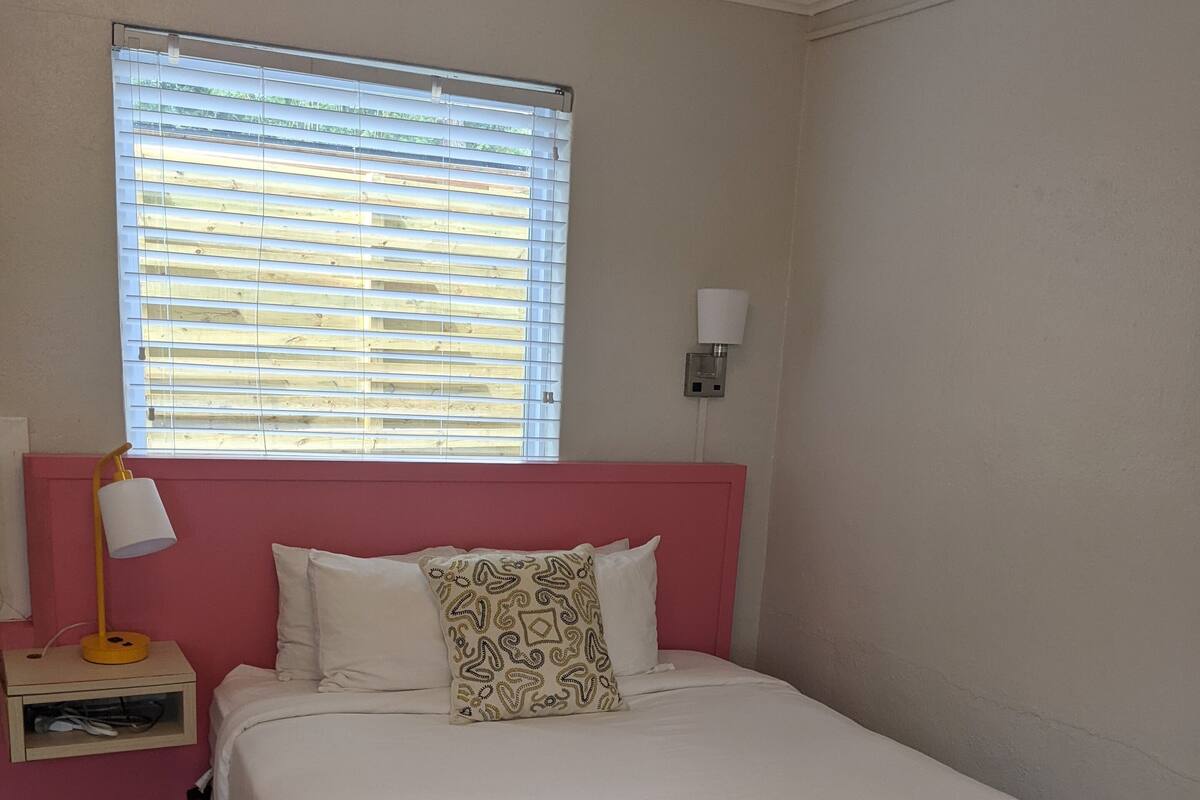 A neatly made bed with white bedding and a patterned pillow, a bedside lamp, and a window with blinds.