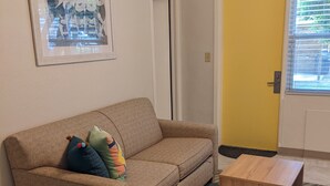 Un salón con un sofá color beige, almohadones coloridos, una mesita de centro de madera, un tapete estampado, un cuadro en la pared y una puerta amarilla.