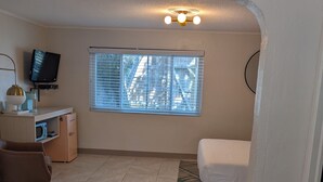 Un espacio habitable pequeño con kitchenette, televisor, ventana con persianas, cama y alfombra estampada.