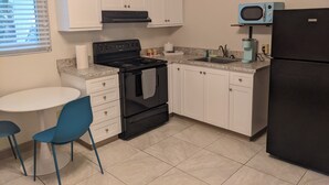 Una cocina con armarios blancos, estufa negra, refrigerador negro, microondas azul, un fregadero y una pequeña mesa con sillas azules.