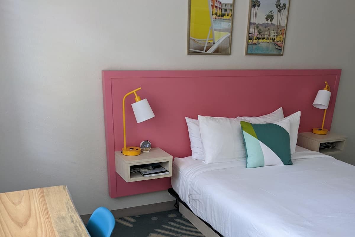 A hotel room with a bed, a desk, and a chair. There are two paintings on the wall and a clock on the bedside table.