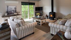 A cozy living room with striped sofas, a fireplace, a television, and a large window with a view of greenery. - French Cottage (Hepburn Springs)