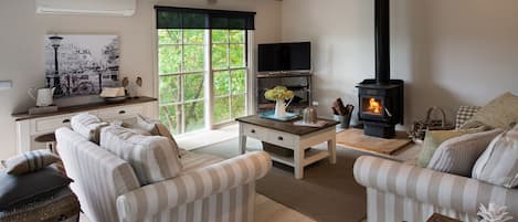 A cozy living room with striped sofas, a fireplace, a television, and a large window with a view of greenery.