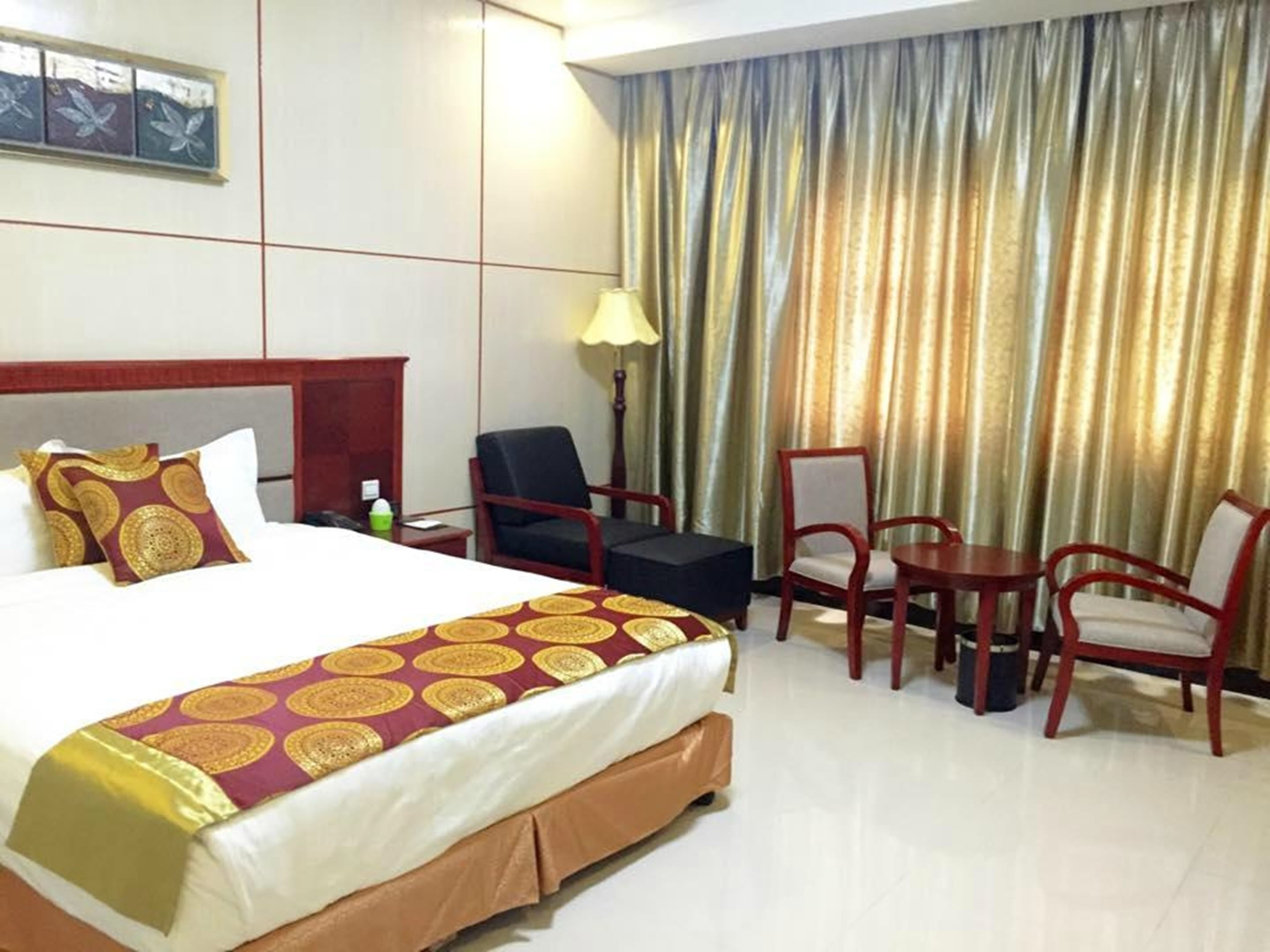 Quarto de hotel com uma cama grande, duas cadeiras, uma mesinha, um abajur e uma cortina.