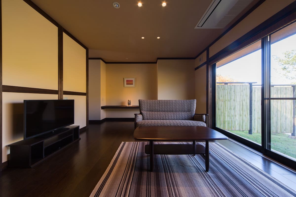 A modern living room with a flat-screen TV, a patterned sofa, and a large window offering a view of the outdoors.