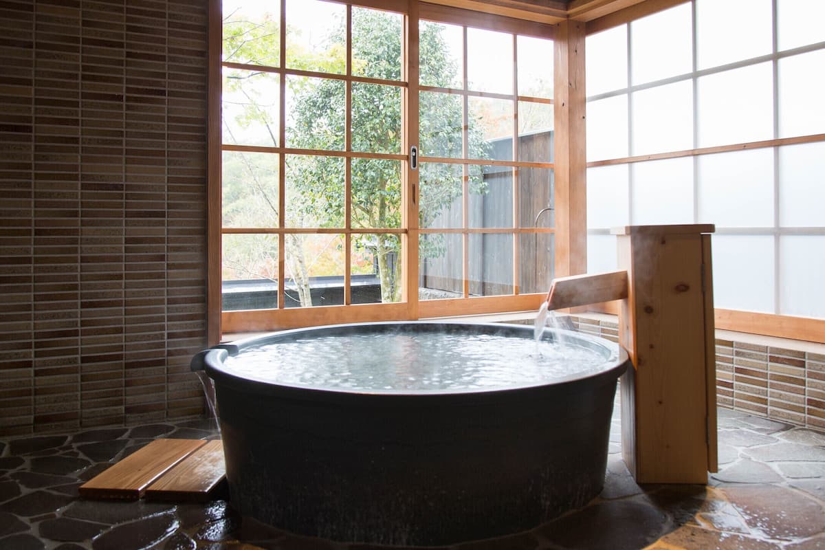 A hot tub in a room with a large window, stone flooring, and wooden walls.