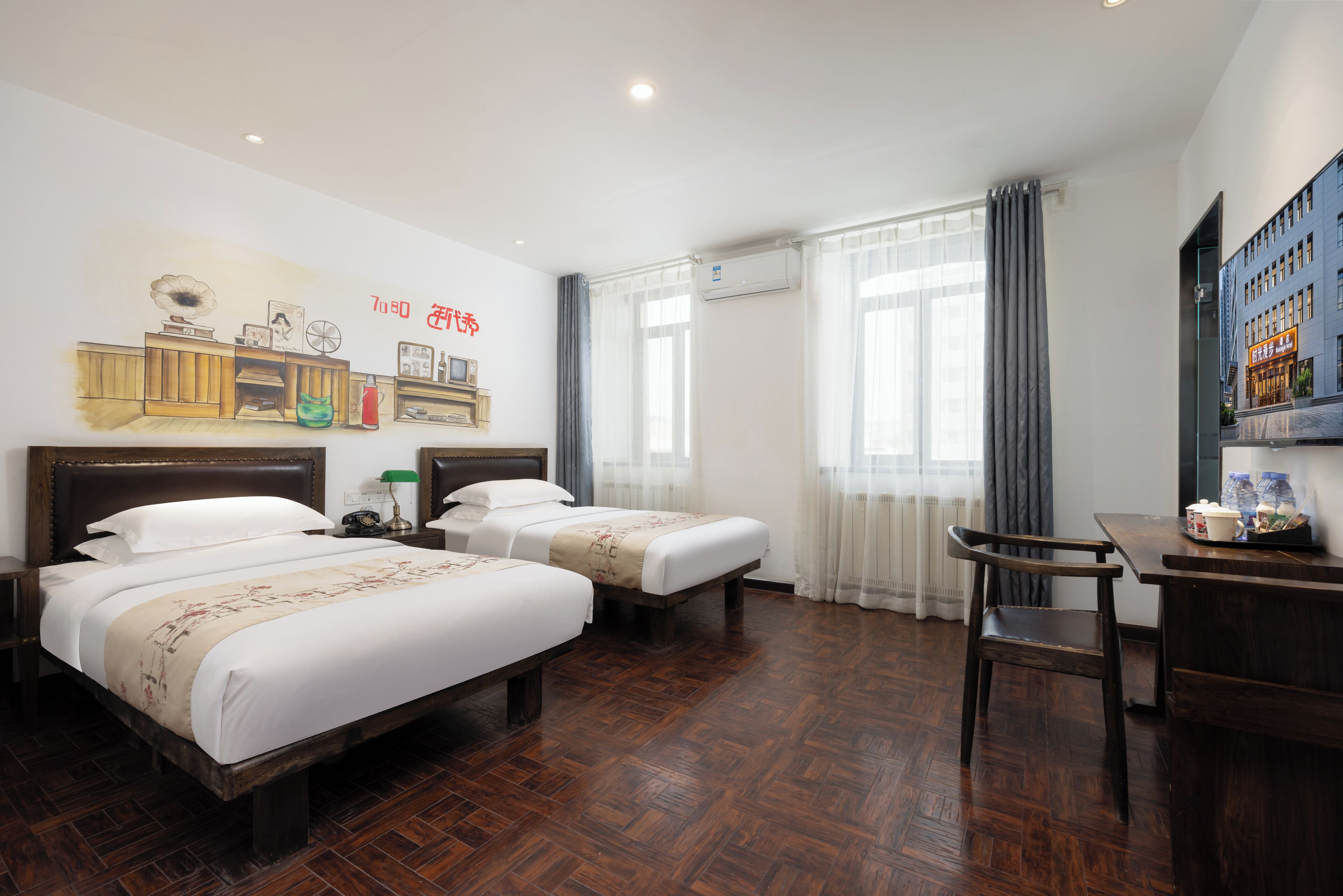 Habitación de hotel con dos camas, un escritorio y una silla. Tiene una ventana con cortinas, un equipo de aire acondicionado y una pared con un mural.