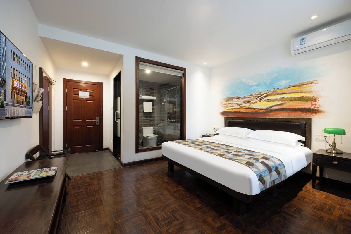 A hotel room with a large bed, a wooden floor, a bathroom with a glass door, and a painting on the wall.