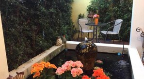 Ein kleines Wasserspiel mit dekorativer Vase und Blumen, umgeben von viel Grün und einer Sitzecke mit Stühlen und Tisch.
