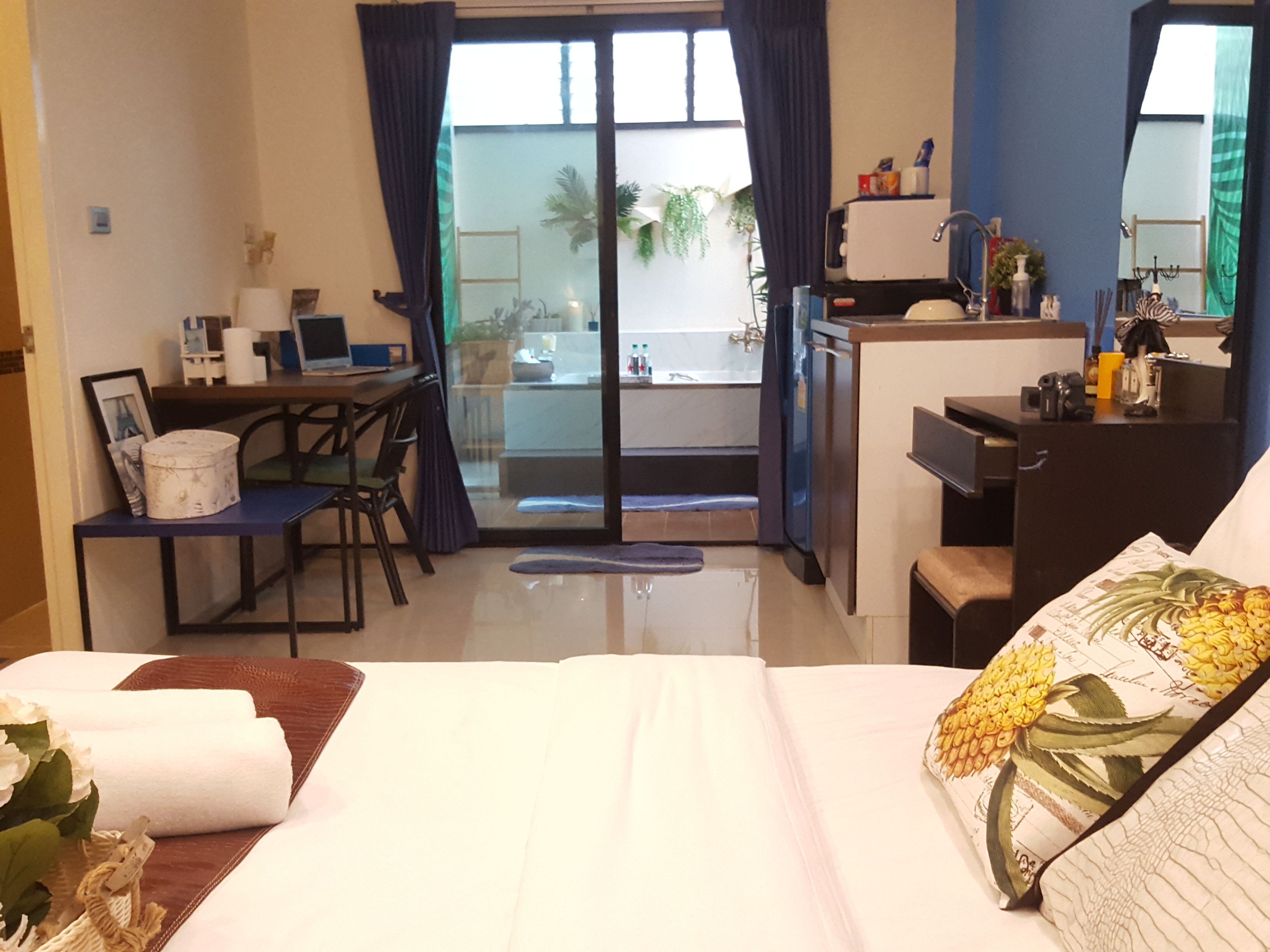 A hotel room with a bed, a desk, a kitchenette, and a balcony with a view of palm trees.