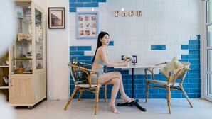 A woman seated at a table in a room with blue and white tiled walls, wicker chairs, and a cabinet with glass doors. - Pace Residence Pattaya (Pattaya)