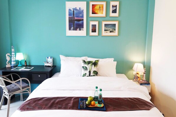 A hotel room with a teal wall, a bed with white bedding, a desk with a lamp, and a tray with fruits and drinks on the bed.