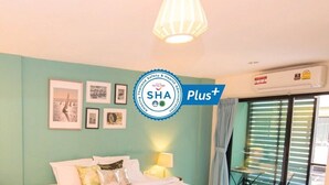 A hotel room with a bed, a nightstand, a lamp, and a window with blinds. There is a sign on the wall and a logo indicating the hotel is part of the SHA Plus+ program. - Pace Residence Pattaya (Pattaya)