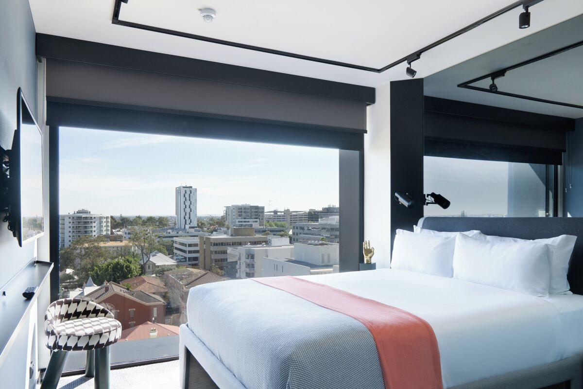 A modern hotel room with a large bed, a view of the city, and a balcony with a chair.