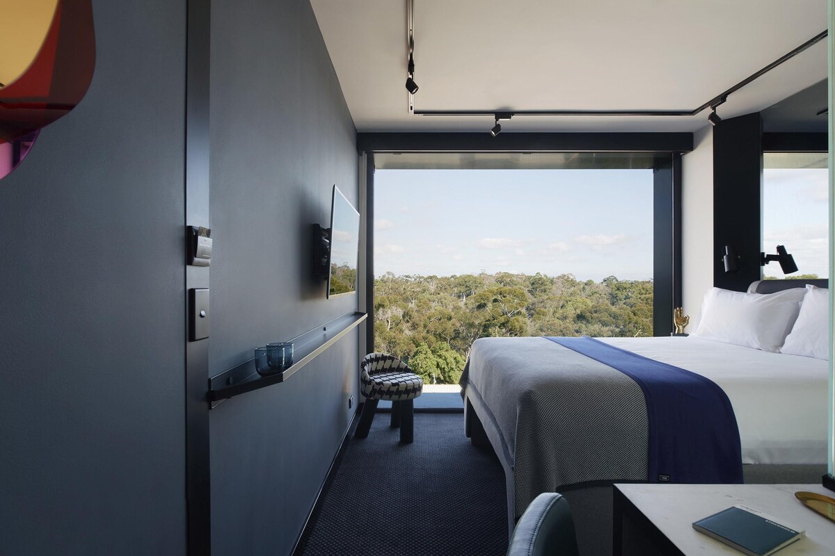 A modern hotel room with a large bed, a desk, and a view of the forest.