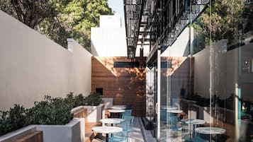 Un patio exterior con mesas y sillas redondas, un banco de madera y una valla metálica.