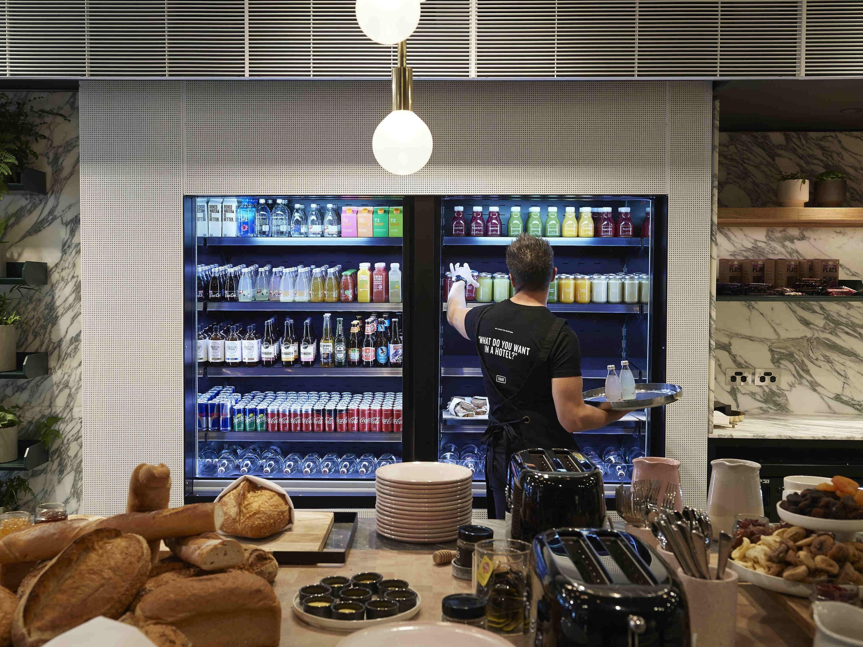 Minibar dell'hotel fornito di bevande e snack, zona buffet con pane e condimenti, e un membro dello staff che serve un vassoio.