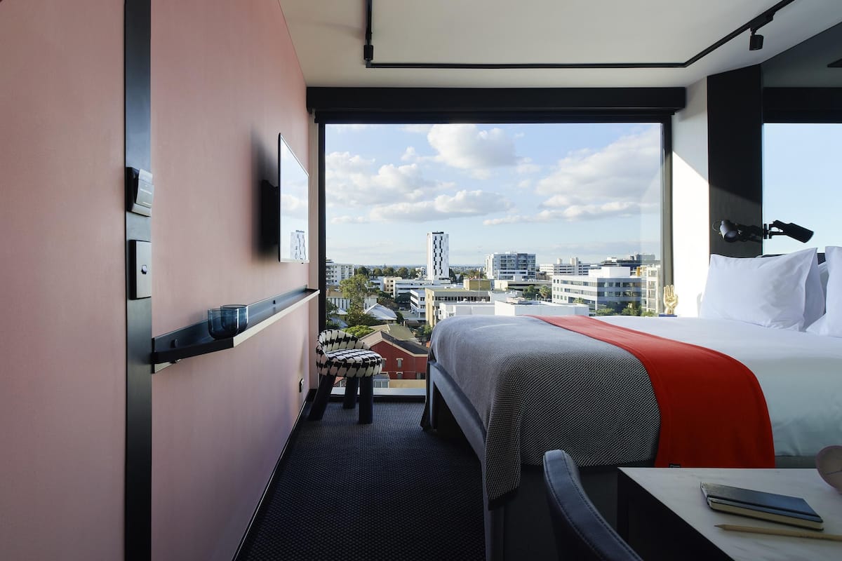 Une chambre d’hôtel comprenant un lit, un bureau, une chaise, une fenêtre donnant sur la ville et un petit tabouret.
