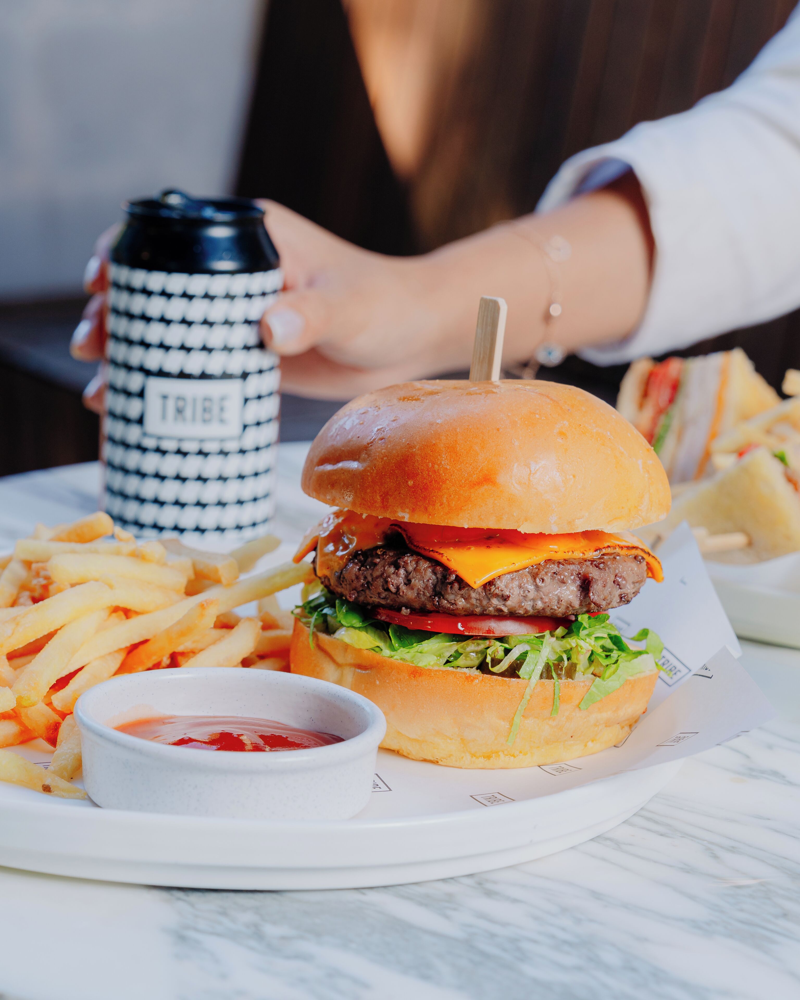 A burger with fries and ketchup on a plate, a drink, and a person in the background.