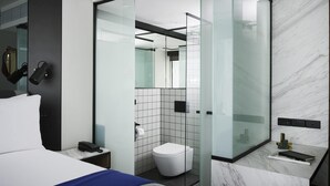 A modern hotel room with a bed, a bedside phone, a glass-enclosed shower, a toilet, and a marble-tiled wall.