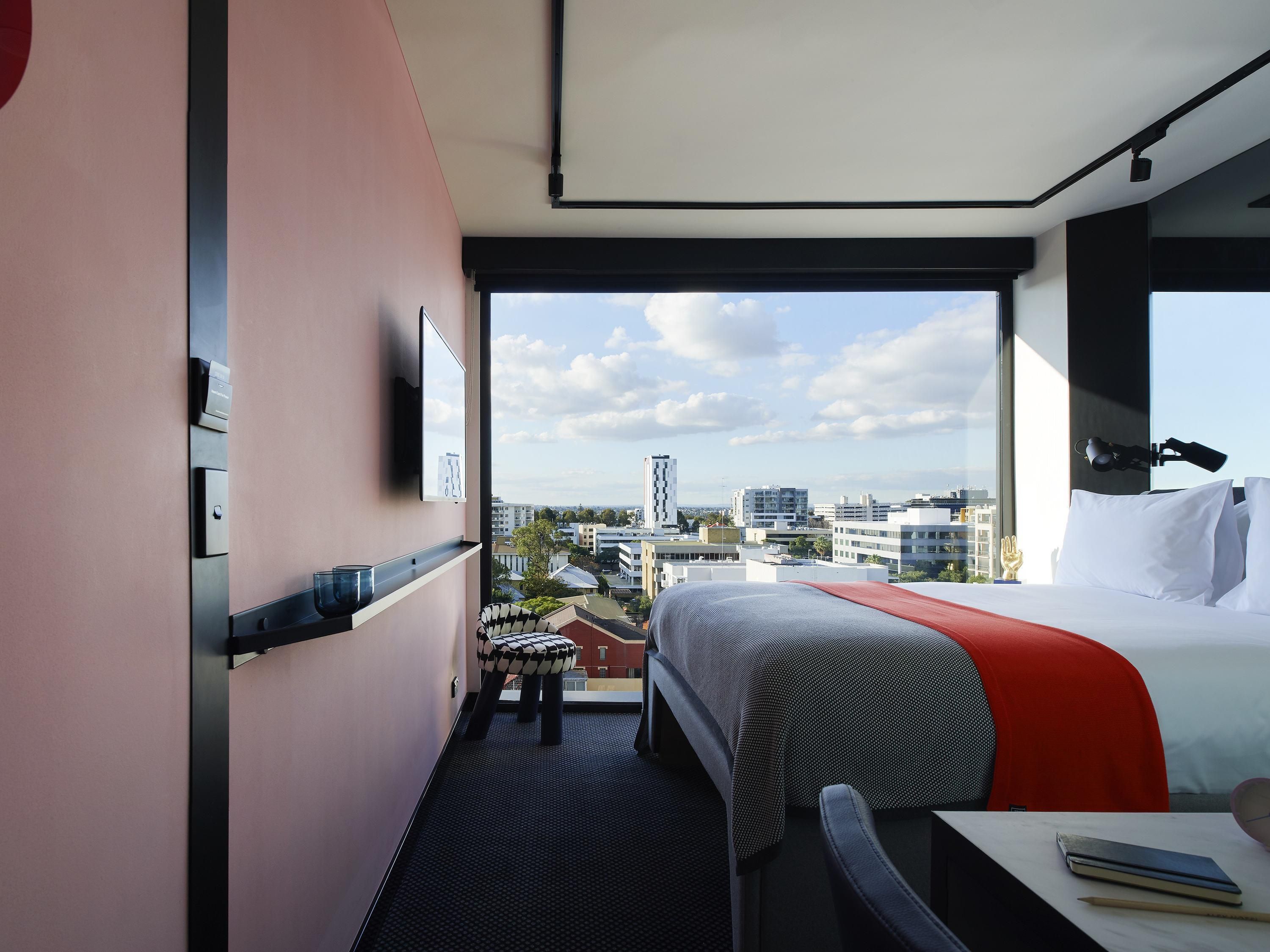 A hotel room with a bed, a desk, a chair, a window with a city view, and a small stool.