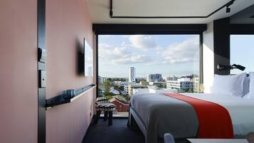 A hotel room with a bed, a desk, a chair, a window with a city view, and a small stool.