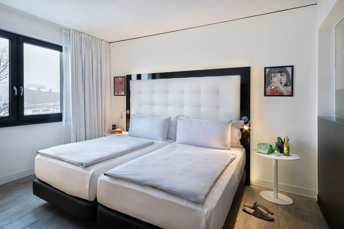 Ein Doppelbett mit einem gepolsterten Kopfteil, ein Nachttisch mit Lampe und Blick auf die Stadt durch ein Fenster mit Vorhang.