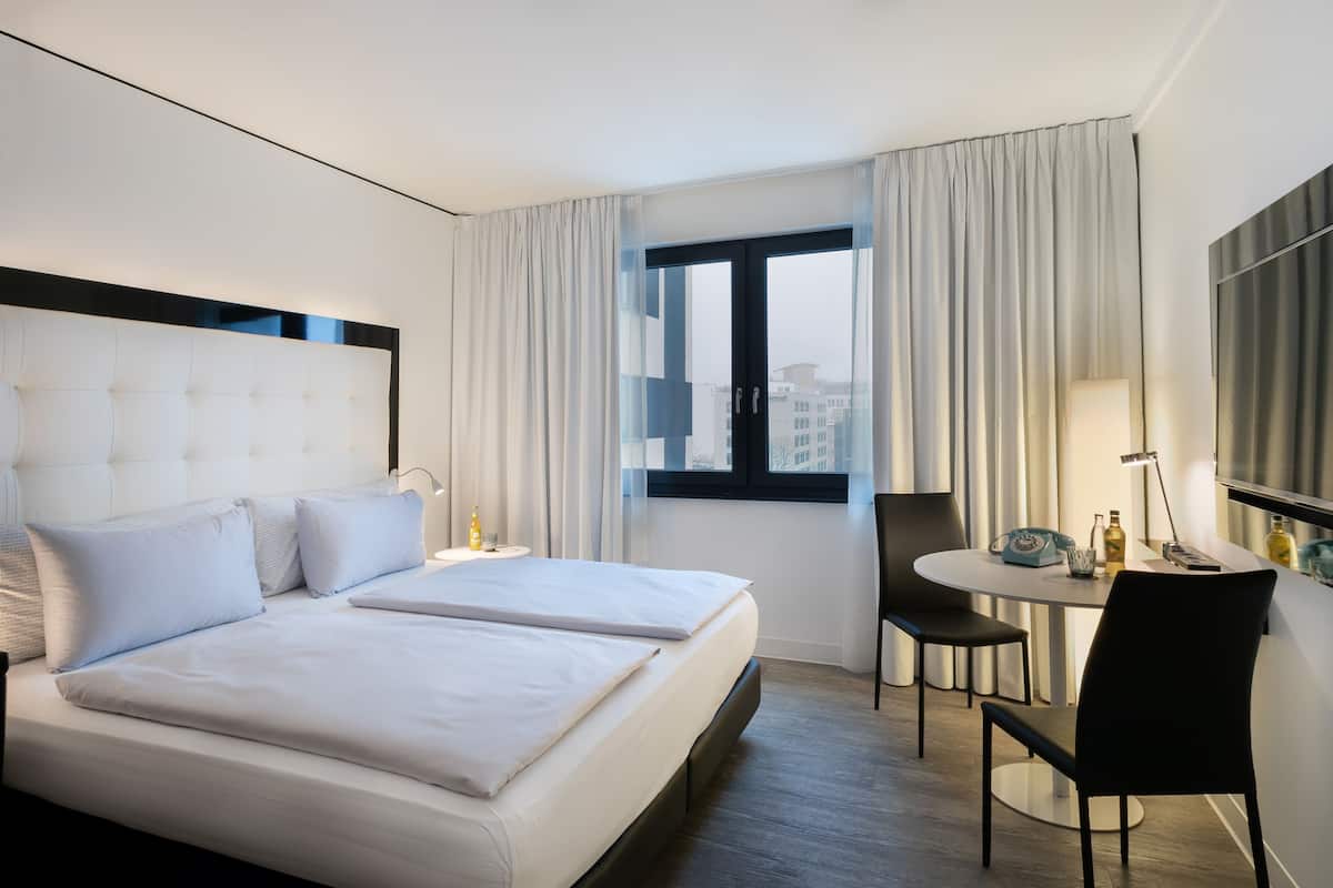 Ein Hotelzimmer mit einem großen Bett, einem kleinen Tisch mit Stuhl, einem Fernseher und einem Fenster mit Vorhängen.