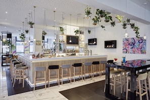 A modern bar area with a long counter, multiple stools, hanging plants, and a large TV screen.