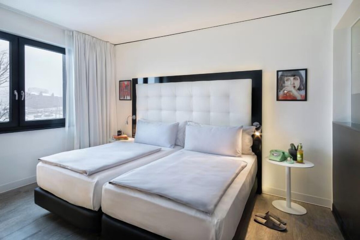 A hotel room with a large bed, a bedside table, a small table with a lamp, and a window with curtains.