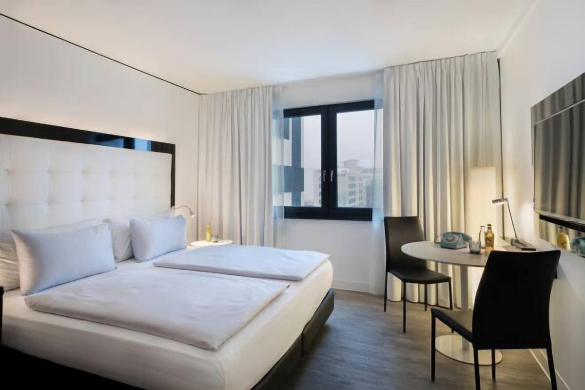 A hotel room with a large bed, a small table with chairs, a window with curtains, and a view of buildings outside.